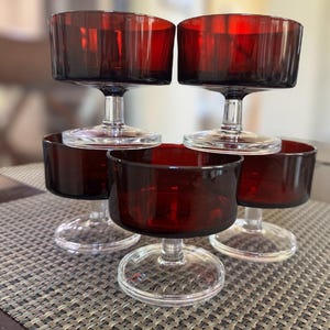 May include: A set of six vintage red glass dessert or cocktail glasses. Each glass features a short, clear stem and a bowl-shaped top with vertical ribbed detailing. The glasses are arranged on a textured surface.