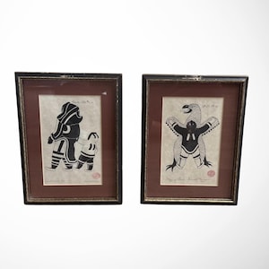 May include: Two framed prints featuring black and white stencil-style illustrations. The left print shows a person in a parka holding a child's hand. The right print depicts a person with arms outstretched beneath a large bird. Both have red seals.