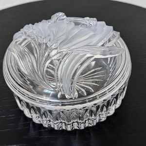 May include: A clear, round glass trinket box with a frosted floral design on the lid. The box has a detailed, textured base and a lid with a raised, three-dimensional flower motif. The box is sitting on a dark surface.