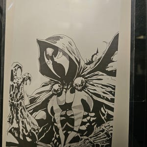 May include: A black and white comic book cover illustration featuring a hooded character with a mask, a flowing cape, and skull-like shoulder accents. The character is depicted with a textured, organic-looking hand reaching forward.