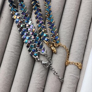 May include: Two beaded bracelets with iridescent blue, green, and purple rectangular beads. One bracelet has a silver clasp and chain, while the other has a gold clasp and chain. The bracelets are displayed on a gray surface.