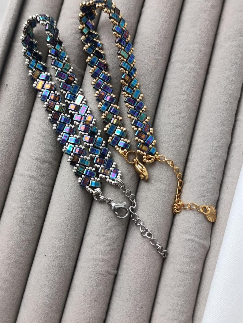 May include: Two beaded bracelets with iridescent blue, green, and purple rectangular beads. One bracelet has a silver clasp and chain, while the other has a gold clasp and chain. The bracelets are displayed on a gray surface.