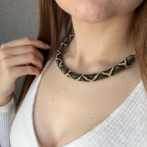 Pode incluir: Um colar de contas com um padrão de cobra. O colar apresenta um design repetitivo de contas pretas e douradas, criando uma aparência texturizada e estampada. O colar é usado ao redor do pescoço.