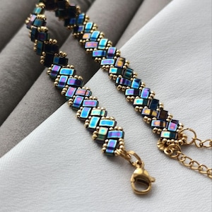 May include: A handmade bracelet featuring iridescent blue and gold beads arranged in a woven pattern. The bracelet has a gold-colored chain and clasp. The jewelry is displayed on a white surface.