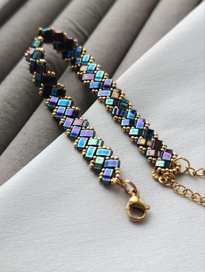 May include: A handmade bracelet featuring iridescent blue and gold beads arranged in a woven pattern. The bracelet has a gold-colored chain and clasp. The jewelry is displayed on a white surface.