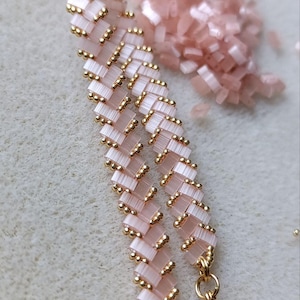 May include: A handmade pink beaded bracelet with gold-colored accents. The bracelet features a herringbone pattern with small, rectangular beads and gold-colored beads. A pile of pink beads is in the background. The bracelet is fastened with a gold-colored clasp.