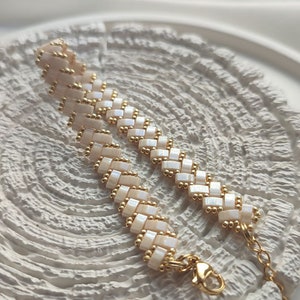 Könnte beinhalten: Ein handgefertigtes Armband mit einem gewebten Muster aus cremefarbenen rechteckigen Perlen und kleinen goldfarbenen Perlen. Das Armband hat eine goldfarbene Kette und einen Verschluss. Der Schmuck wird auf einer weißen, strukturierten Oberfläche präsentiert.
