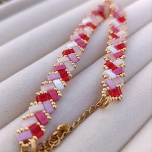May include: A beaded bracelet featuring a herringbone pattern. The bracelet is made with small, rectangular beads in shades of red, pink, and white, accented with gold-colored beads. A delicate gold chain and clasp complete the design.