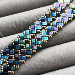 May include: Close-up of a beaded bracelet with an iridescent, rectangular design. The beads shift in color from blue to purple and gold. The bracelet is set against a gray, textured background.