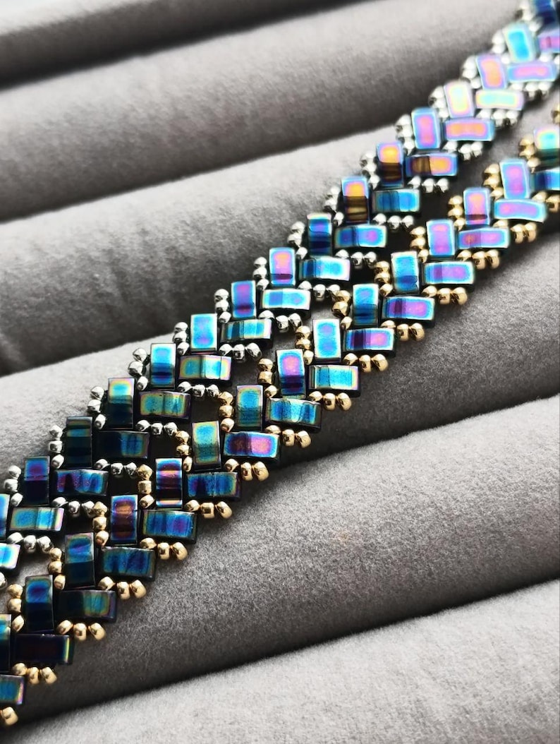 May include: Close-up of a beaded bracelet with an iridescent, rectangular design. The beads shift in color from blue to purple and gold. The bracelet is set against a gray, textured background.