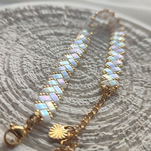 May include: A delicate bracelet featuring iridescent white rectangular beads woven together with gold-colored accents. The bracelet has a gold-colored chain and clasp, and a small sun-shaped charm. The jewelry is displayed on a textured white surface.
