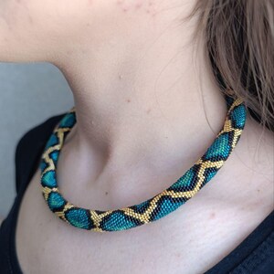 Puede incluir: Un collar tubular de cuentas con un patrón geométrico repetitivo de cuentas de vidrio en color verde azulado, negro y dorado. El collar tiene una forma cilíndrica y se ajusta al cuello, mostrando una textura consistente de cuentas pequeñas.