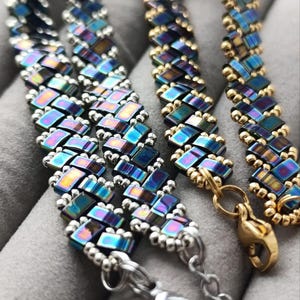 May include: Close-up of three beaded bracelets. The bracelets feature iridescent, rectangular beads in shades of blue, purple, and gold. The bracelets have silver and gold clasps. The beads are arranged in a repeating pattern.