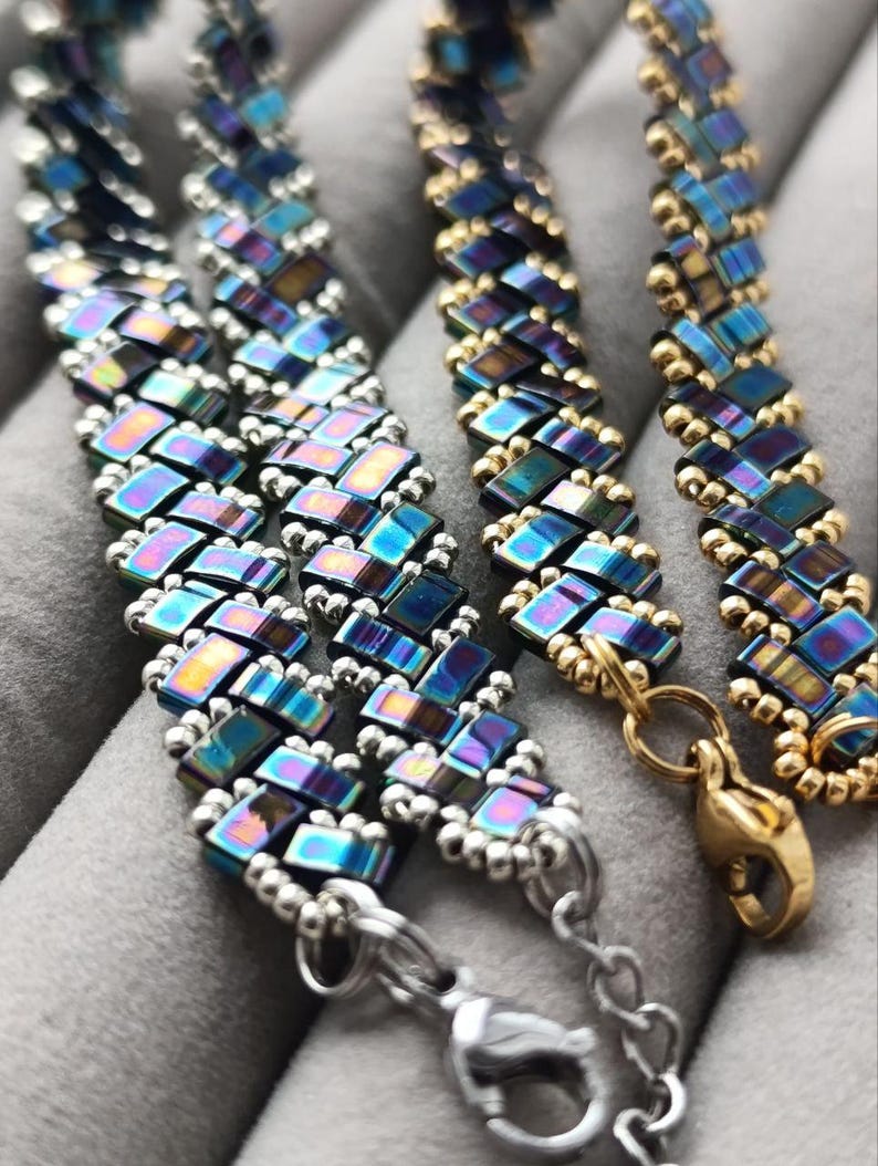 May include: Close-up of three beaded bracelets. The bracelets feature iridescent, rectangular beads in shades of blue, purple, and gold. The bracelets have silver and gold clasps. The beads are arranged in a repeating pattern.