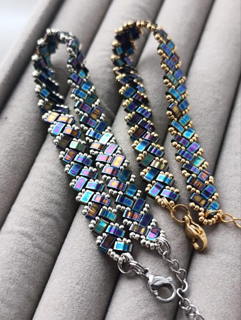 May include: Two beaded bracelets with iridescent blue, purple, and gold accents. One bracelet has silver-toned clasps and chain, while the other has gold-toned clasps. The bracelets are made of small, rectangular beads.