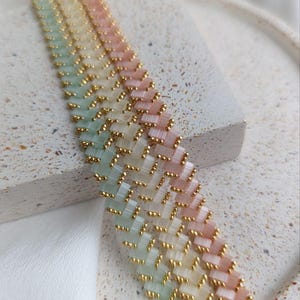 Peut inclure: Un bracelet de perles aux couleurs pastel avec un motif en chevrons. Le bracelet présente un dégradé de couleurs, dont le vert clair, le jaune et le rose, avec des perles dorées. Le bracelet est présenté sur une surface texturée.