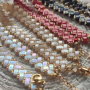 May include: Close-up of several beaded bracelets in various colors, including red, white, black, and iridescent. The bracelets feature a woven design with gold-colored accents and clasps. The bracelets are displayed on a textured surface.