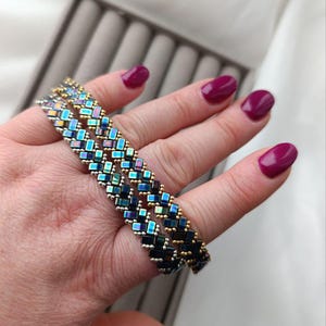 May include: A beaded bracelet with a geometric pattern. The bracelet features iridescent blue, green, and gold beads. The bracelet is shown on a hand with polished nails.
