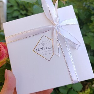 May include: A white gift box tied with a white ribbon with silver trim. The box has a gold and white logo that reads "JEWELLE Fashion Style". The background includes green foliage and a pink and yellow rose.