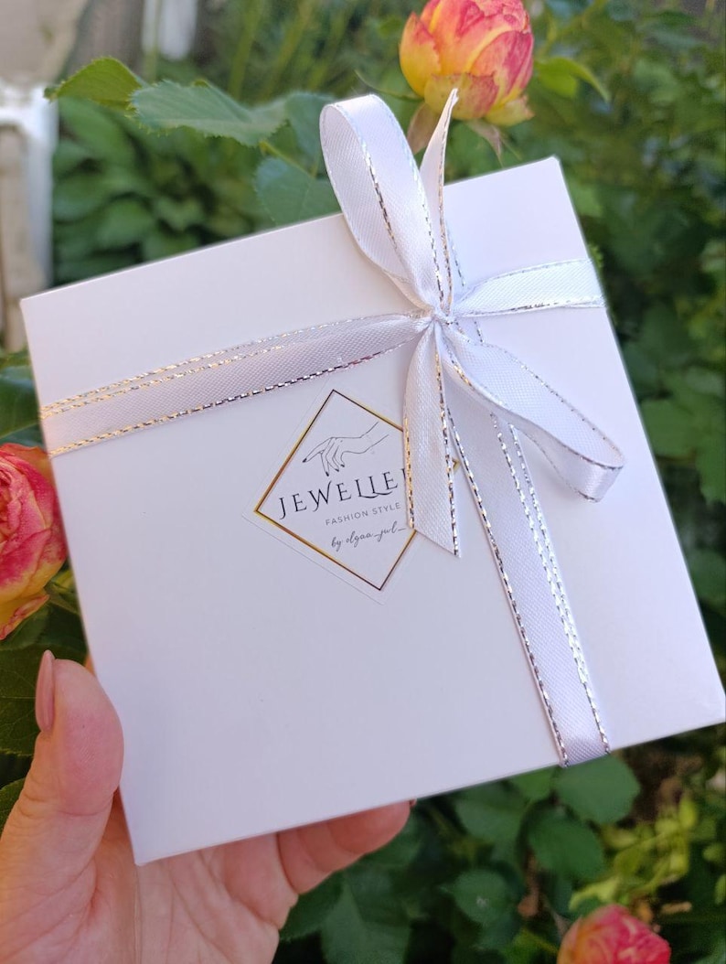 May include: A white gift box tied with a white ribbon with silver trim. The box has a gold and white logo that reads "JEWELLE Fashion Style". The background includes green foliage and a pink and yellow rose.