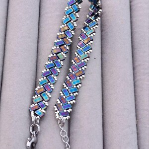 May include: A beaded bracelet with a herringbone pattern. The beads are iridescent, displaying shades of blue, purple, and gold. The bracelet has a silver-toned clasp and chain extender. The jewelry is displayed on a gray surface.