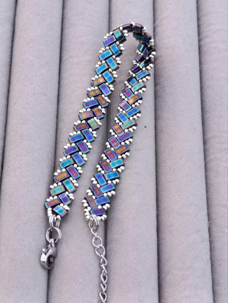 May include: A beaded bracelet with a herringbone pattern. The beads are iridescent, displaying shades of blue, purple, and gold. The bracelet has a silver-toned clasp and chain extender. The jewelry is displayed on a gray surface.