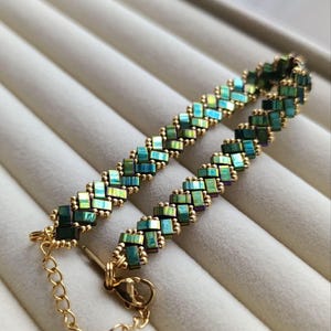 May include: A beaded bracelet with a geometric pattern. The bracelet features iridescent green and blue beads, interspersed with gold-colored beads. The clasp and chain are also gold-colored. The bracelet is displayed on a light gray surface.