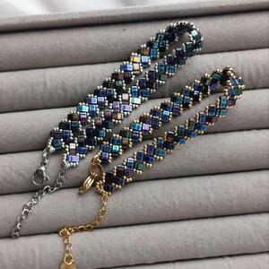 May include: Two beaded bracelets with iridescent square beads in shades of blue, green, and purple. One bracelet has a silver clasp and chain, while the other has a gold clasp and chain. The bracelets are displayed on a gray surface.