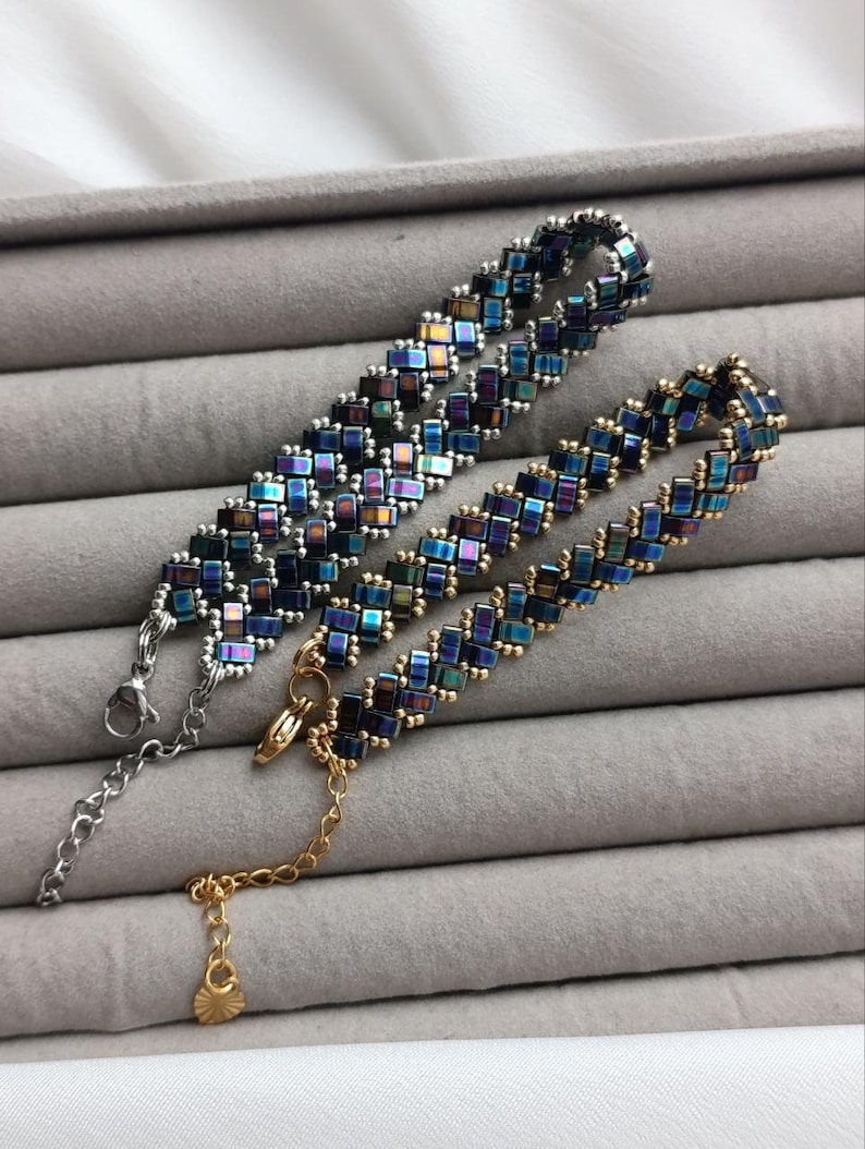 May include: Two beaded bracelets with iridescent square beads in shades of blue, green, and purple. One bracelet has a silver clasp and chain, while the other has a gold clasp and chain. The bracelets are displayed on a gray surface.