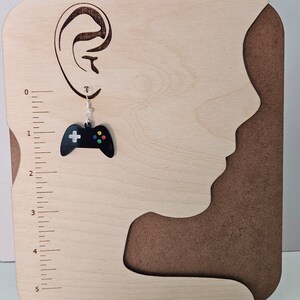 May include: A black video game controller earring with a silver hook. The controller has a white plus sign and colorful buttons. The earring is displayed on a wooden background with a ruler and a cutout of a human profile.
