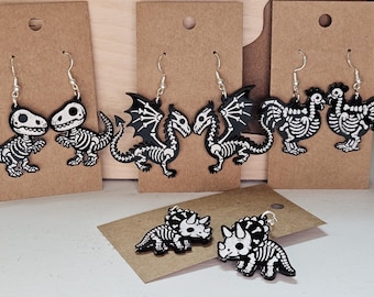 Puede incluir: Pendientes y colgante de esqueleto de dinosaurio, dragón y pollo en blanco y negro. Los pendientes se exhiben en cartulina marrón. El colgante es un esqueleto de triceratops. Los pendientes tienen ganchos plateados.