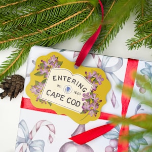 May include: A gold-coloured gift tag with a scalloped edge, featuring a floral border of purple flowers and the text "ENTERING CAPE COD" with "INC. 1620" and a small blue emblem in the centre. The tag is attached by a red ribbon to a gift.