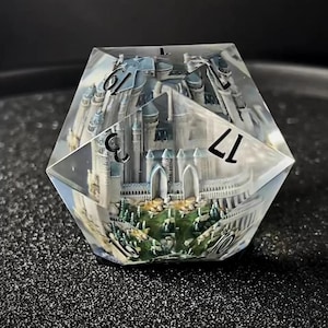May include: A clear, multi-sided die with a miniature castle scene inside. The castle features grey towers, green trees, and a grassy landscape. The die has white numbers on its faces, including the number 17.