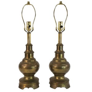 May include: A pair of matching brass table lamps featuring a rounded, bulbous base with a stepped pedestal design. Each lamp includes a vertical brass neck, a standard light bulb socket with a black switch, and a wire harp frame for a lampshade.