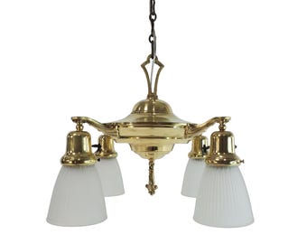 May include: A polished brass chandelier featuring a central tiered body with four downward-facing arms. Each arm holds a bell-shaped, ribbed, frosted glass shade. The fixture is suspended by a dark metal chain and includes a decorative finial.