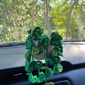 May include: A crocheted green plant hanging from a rearview mirror in a car. The plant is in a white crocheted pot and has long green vines.