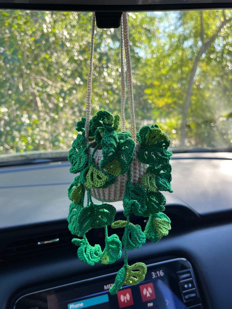 May include: A crocheted green plant hanging from a rearview mirror in a car. The plant is in a white crocheted pot and has long green vines.