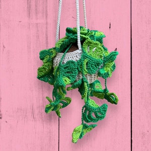May include: A crocheted hanging planter with green leaves shaped like monstera plants. The planter is hanging from a white cord against a pink wooden background.
