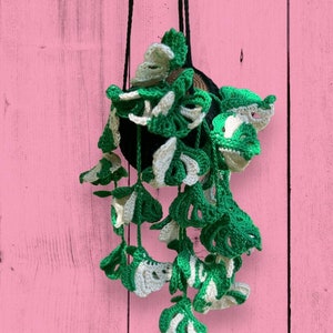 May include: A crocheted hanging planter with green and white leaves. The planter is hanging from a black cord against a pink wooden background.