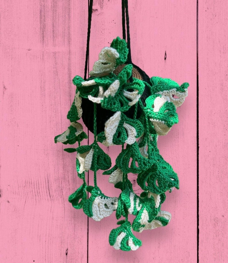 May include: A crocheted hanging planter with green and white leaves. The planter is hanging from a black cord against a pink wooden background.