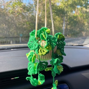 May include: A crocheted green plant hanging from a rearview mirror in a car. The plant is in a cream-colored pot and has long, trailing vines.