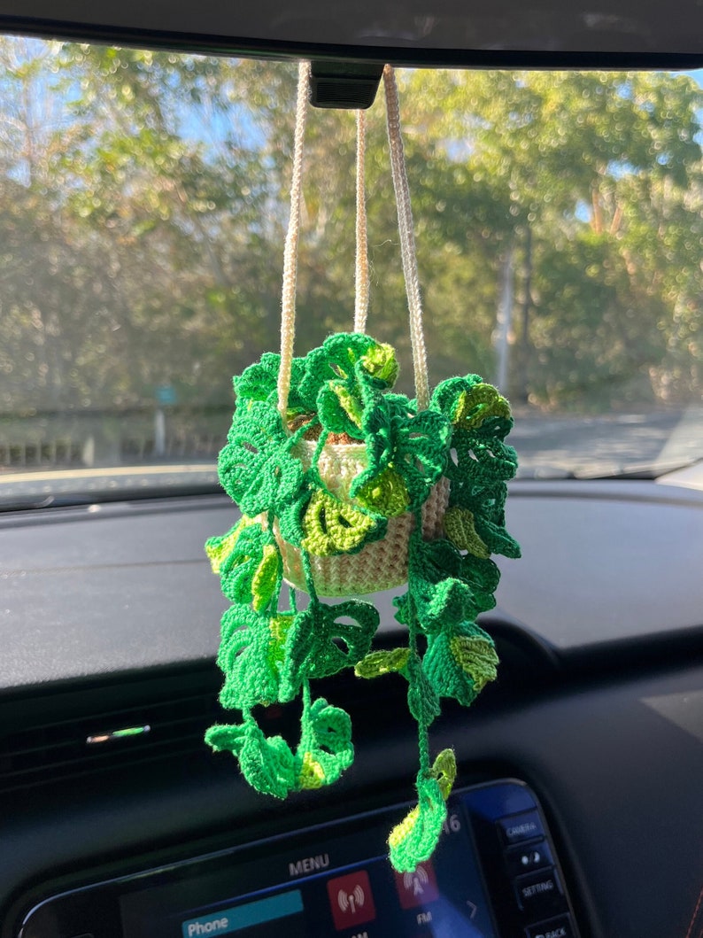 May include: A crocheted green plant hanging from a rearview mirror in a car. The plant is in a cream-colored pot and has long, trailing vines.