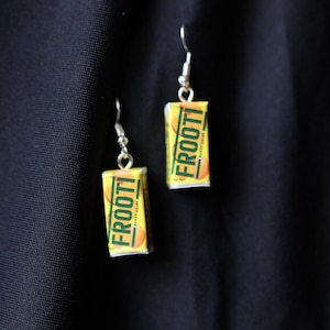 May include: A pair of silver earrings with a yellow and green Frootti juice box charm on each earring. The juice box charm has the words 