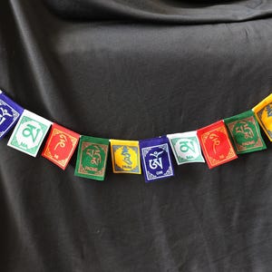 May include: A string of colorful prayer flags with various symbols and text in white, green, and yellow. The flags are rectangular and attached to a white string. The background is a dark, solid color.