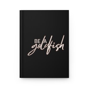 May include: Black journal with the words "BE A goldfish" in a cursive font. The text is in a light brown color with a white outline. The journal is closed and has a smooth, matte finish.