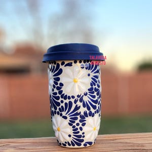 May include: A white ceramic travel mug with a blue and white floral pattern. The mug has a blue silicone lid and the text "Talavera Artistico" is visible on the side.
