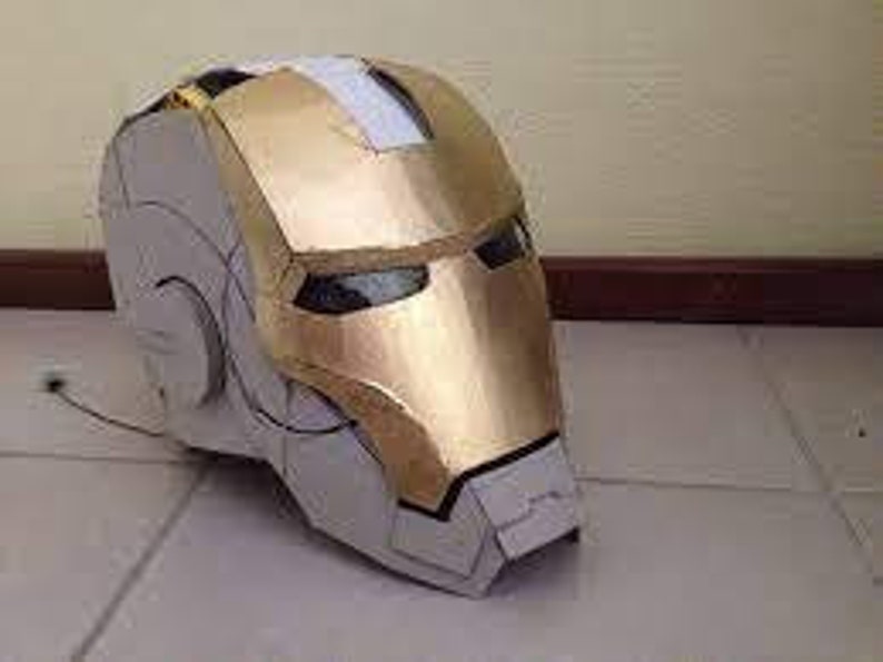 May include: A gold and white cardboard Iron Man helmet. The helmet has a detailed design with a gold faceplate and a white interior.