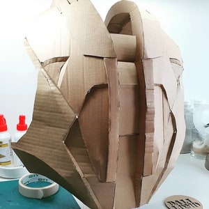 May include: A brown cardboard sculpture of a helmet or headpiece. The sculpture is made of many pieces of cardboard that have been cut and glued together. The sculpture is sitting on a table with other crafting supplies.