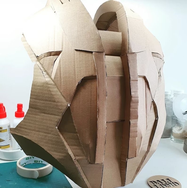 May include: A brown cardboard sculpture of a helmet or headpiece. The sculpture is made of many pieces of cardboard that have been cut and glued together. The sculpture is sitting on a table with other crafting supplies.