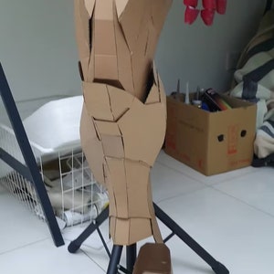 May include: A cardboard model of a robot leg, painted red and brown, with a black stand. The leg is made of multiple cardboard pieces, and the foot is separate.